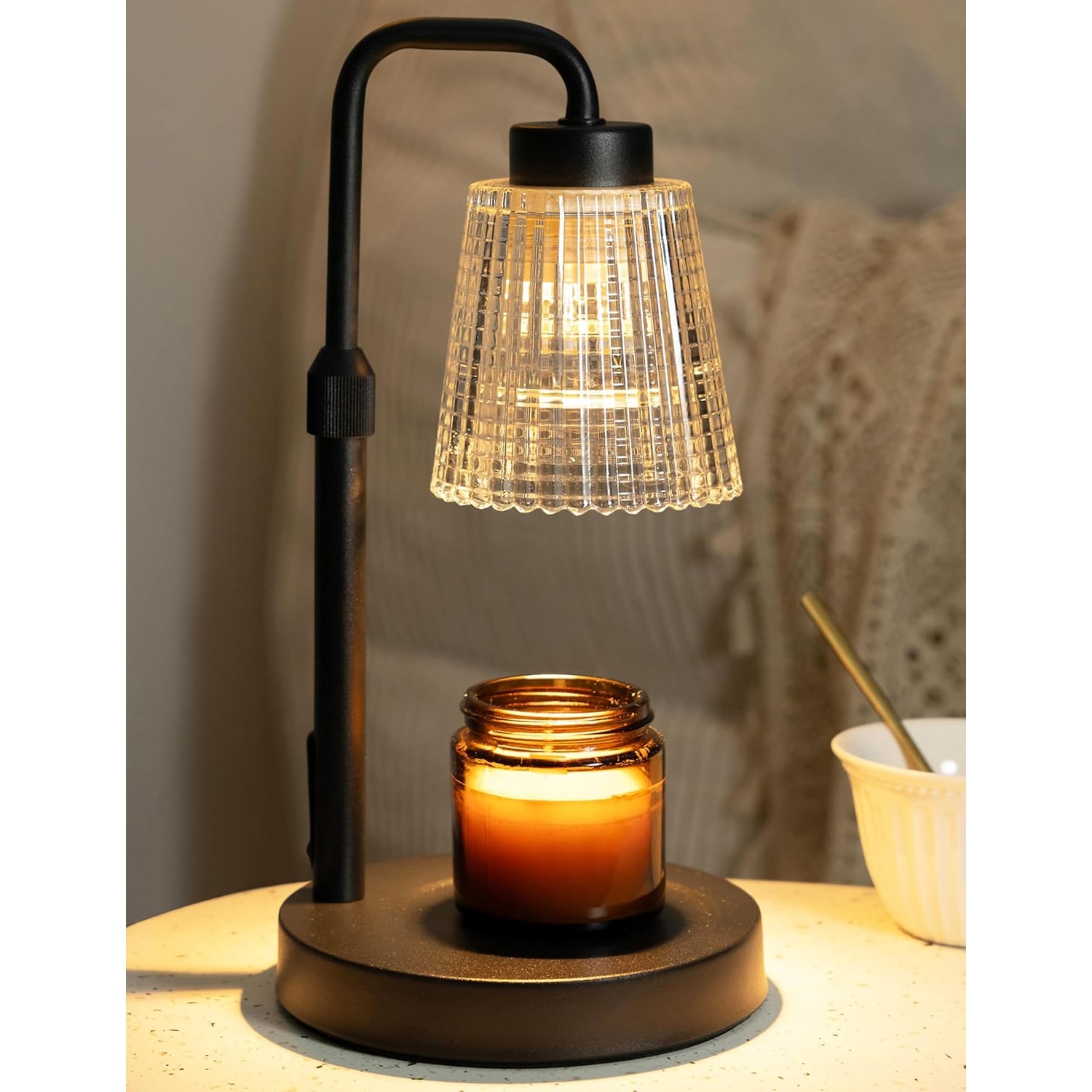 Matt black candle warmer lamp with a fluted glass shade glowing over an amber jar candle on a bedside table