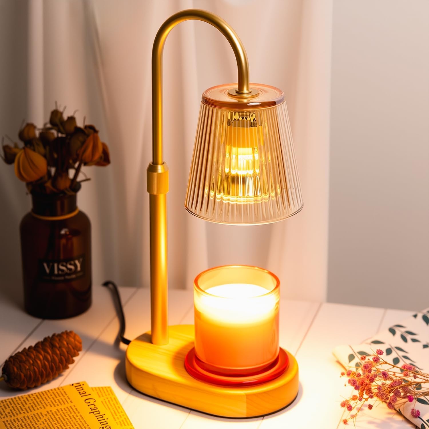Amber glass candle warmer lamp lit above a glowing jar candle on a wooden table beside dried flowers
